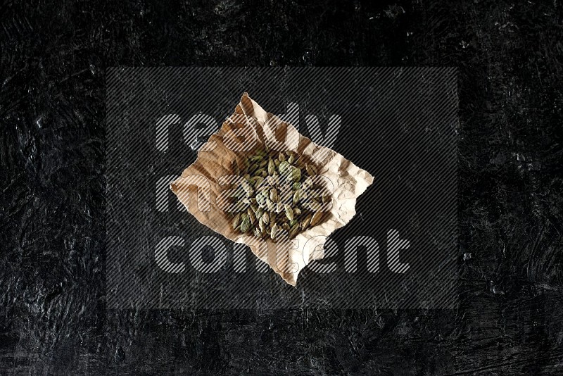 Cardamom seeds in a crumpled piece of paper on textured black flooring