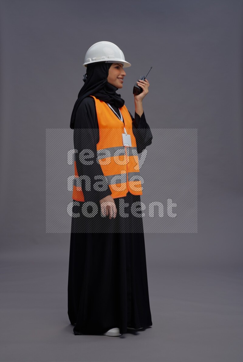 Saudi woman wearing Abaya with engineer vest with neck strap employee badge standing holding walkie-talkie on gray background