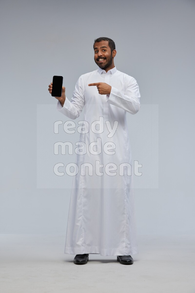 Saudi man wearing Thob standing showing phone to camera on Gray background
