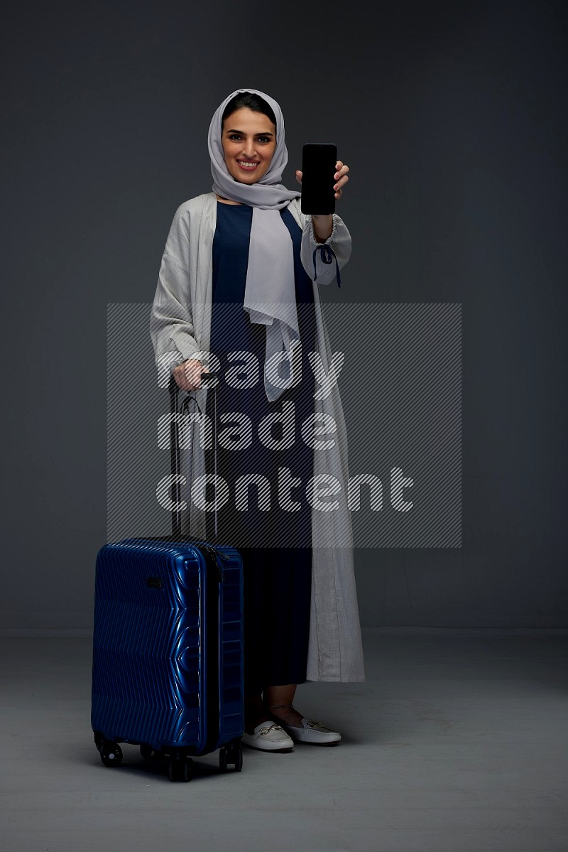 A Saudi woman wearing a light gray Abaya and head scarf standing and showing the phone's screen while wearing headphones eye level on a grey background