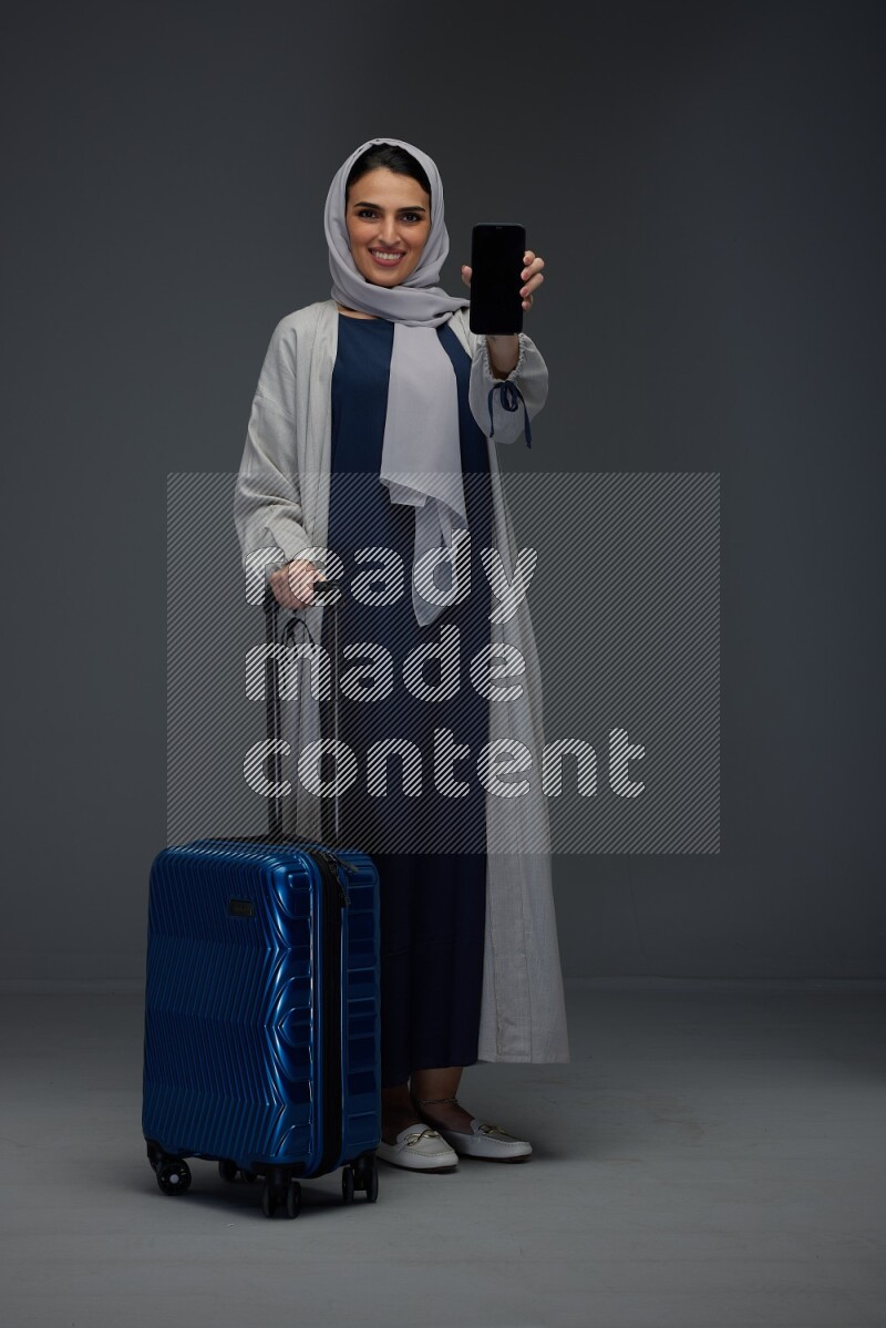 A Saudi woman wearing a light gray Abaya and head scarf standing and showing the phone's screen while wearing headphones eye level on a grey background