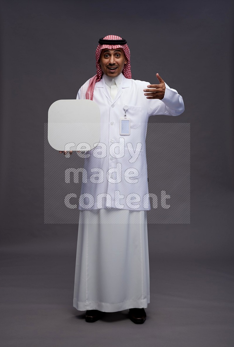 Saudi man wearing thob with lab coat and shomag with pocket employee badge standing holding social media sign on gray background