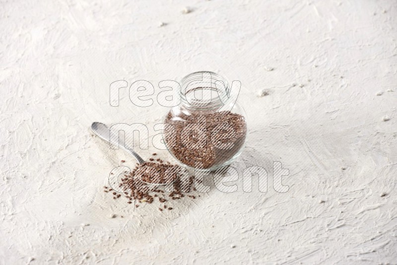 A glass spice jar full of flax and a metal spoon full of the seeds on a textured white flooring in different angles