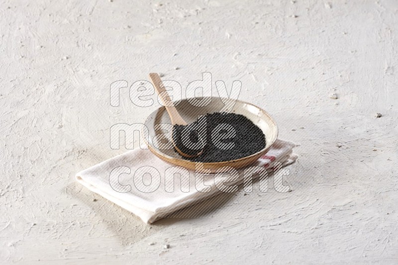 A multicolored pottery plate full of black seeds with a wooden spoon full of the seeds on a napkin on a textured white flooring