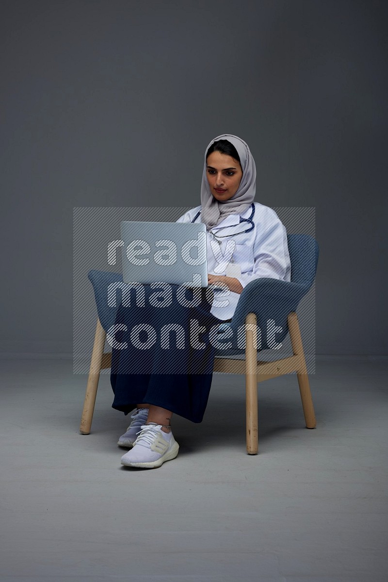 A doctor wearing a light gray head scarf sitting on a dark grey chair and using a laptop eye level on a grey background