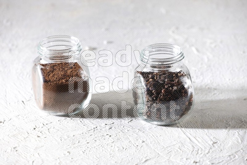2 glass jars full of cloves powder and cloves grains on a textured white flooring