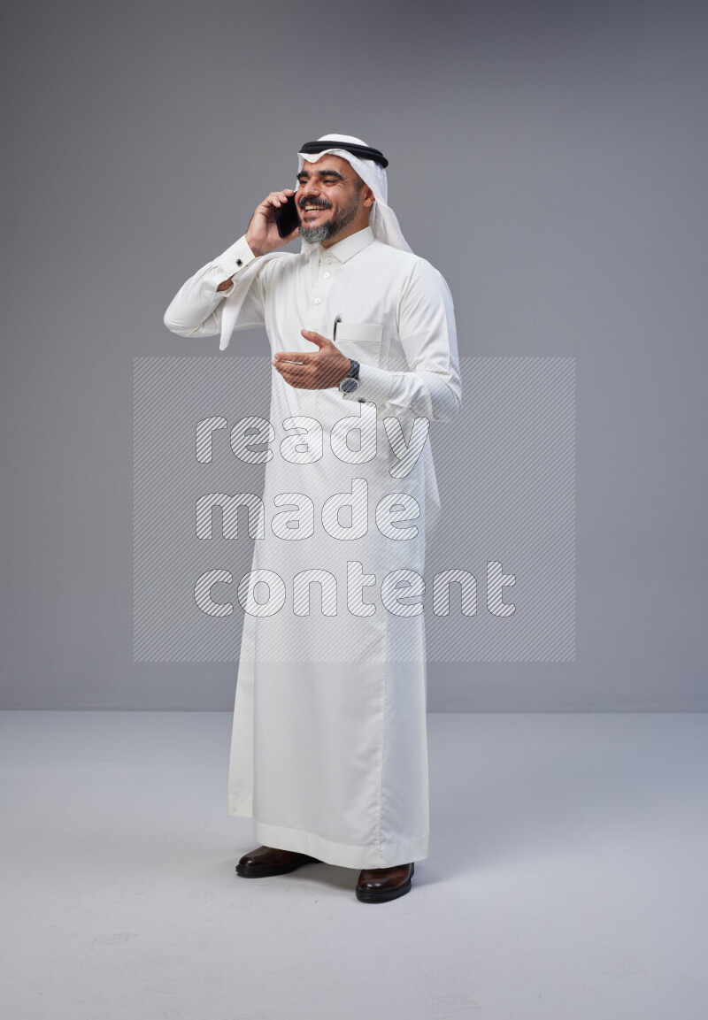 Saudi man Wearing Thob and white Shomag standing talking on phone on Gray background