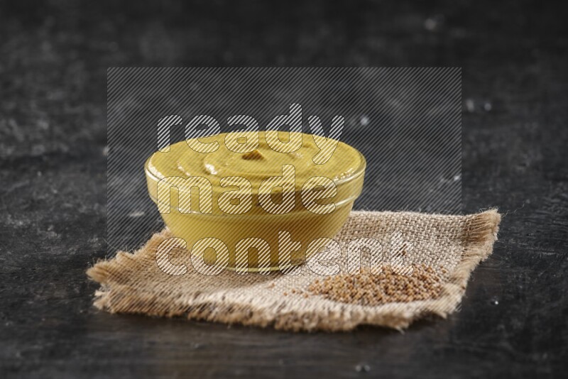 A glass bowl full of mustard paste set on a burlap piece with some mustard seeds on a textured black flooring