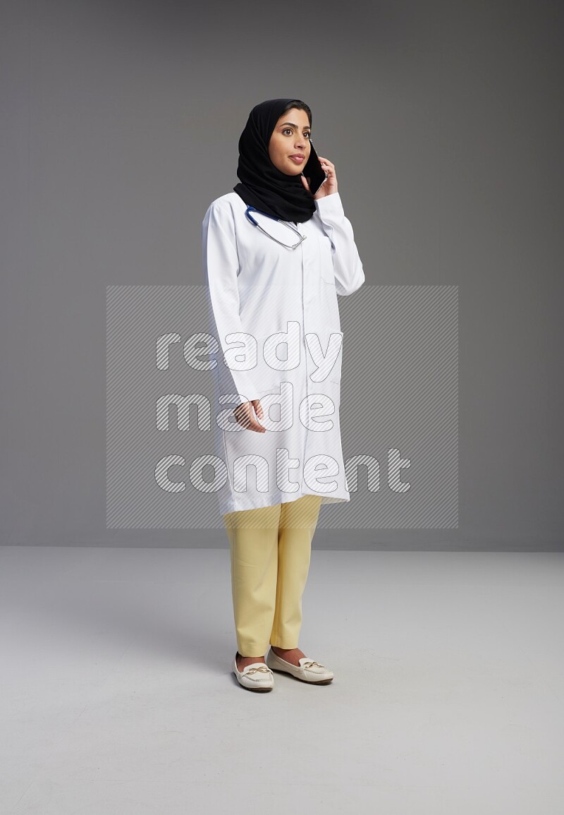 Saudi woman wearing lab coat with stethoscope standing talking on phone on Gray background