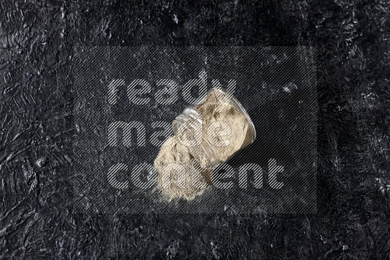 A flipped glass jar full of white pepper powder with spilled powder on textured black flooring