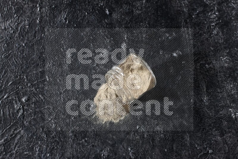 A flipped glass jar full of white pepper powder with spilled powder on textured black flooring