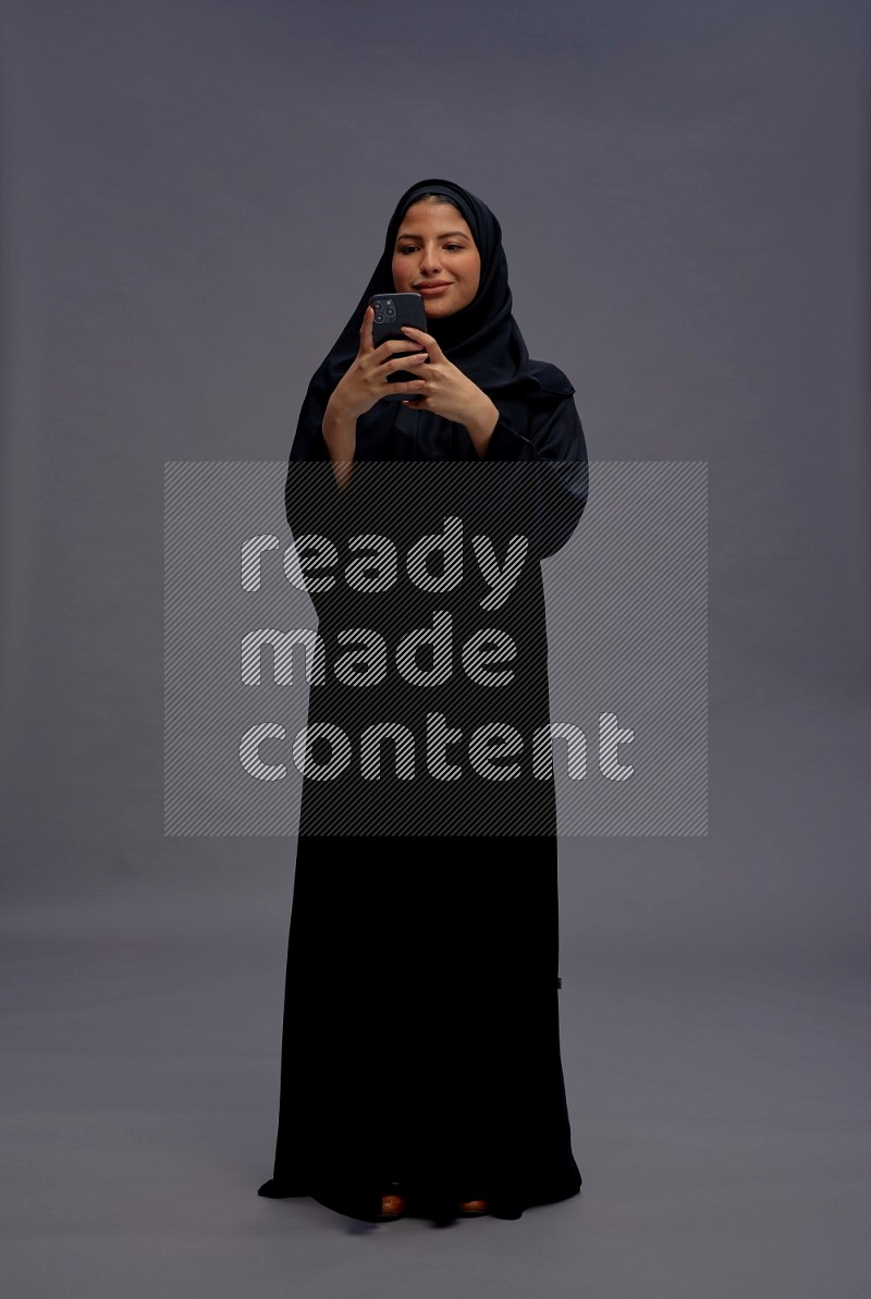 Saudi woman wearing Abaya standing texting on phone on gray background