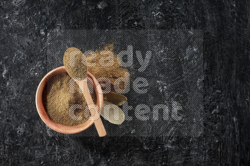 A wooden bowl and spoon full of cumin powder on a textured black flooring