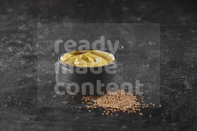 A black pottery bowl full of mustard paste with mustard seeds underneath on textured black flooring
