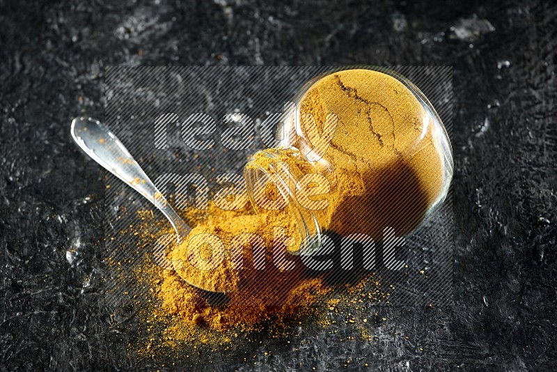 A flipped glass spice jar and a metal spoon full of turmeric powder and powder spilled out of it on textured black flooring