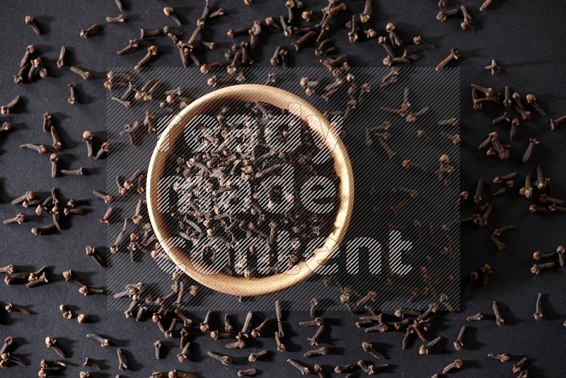 A wooden bowl full of cloves with spreaded whole cloves on a black flooring