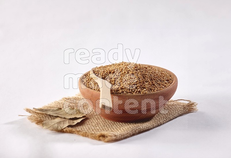 A wooden bowl full of mustard seeds with a wooden spoon on the seed set on burlap fabric on a white flooring