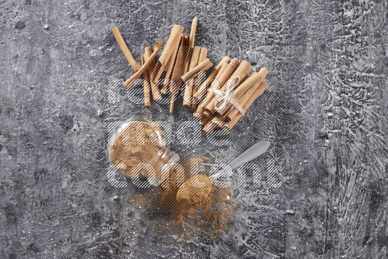 Herbal glass jar full cinnamon powder flipped and a metal spoon full of powder, cinnamon sticks stacked and bounded in the back on textured black background in different angles