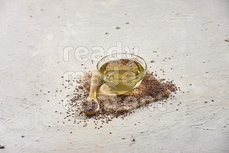 A glass bowl full of flaxseeds oil and wooden spoon full of flaxseeds with spread seeds on burlap fabric on a textured white flooring