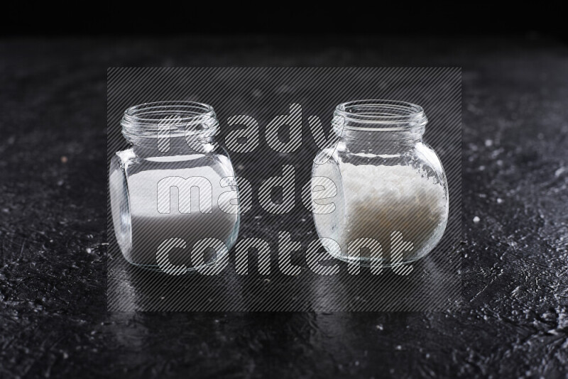 2 glass jars one is filled with table salt and the other with coarse sea salt on black background