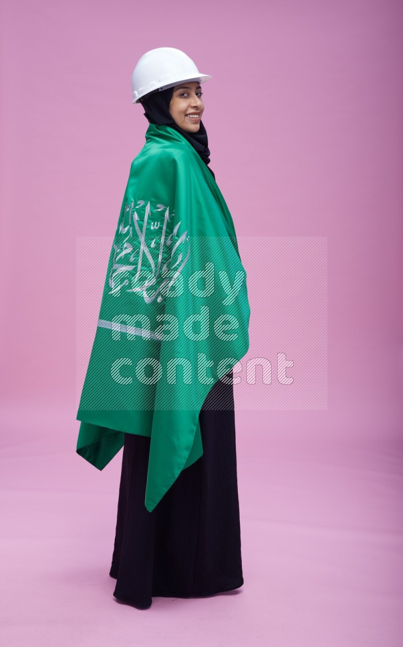 Saudi woman wearing Abaya with engineer vest and helmet standing holding Saudi flag on pink background