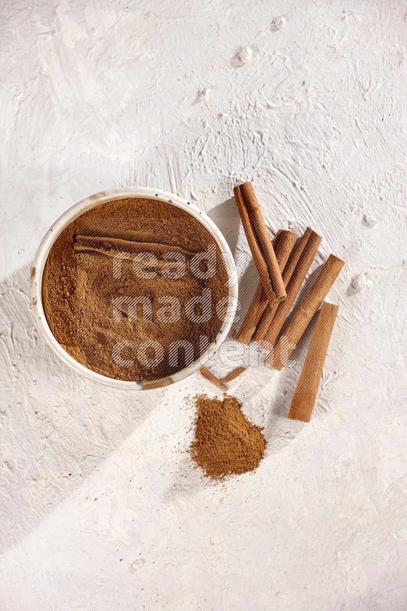 Ceramic bowl full of cinnamon powder with cinnamon sticks on the side on white background