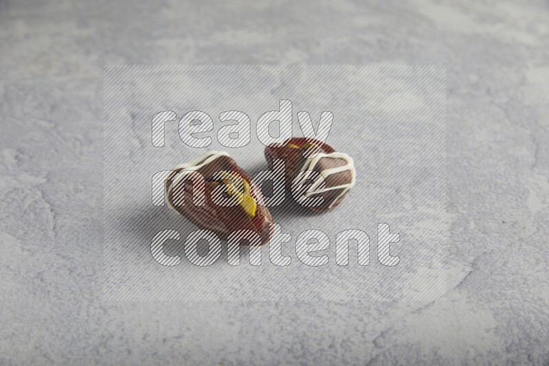 two pistachio stuffed dates covered with dark chocolate with white chocolate drizzle on a light grey background