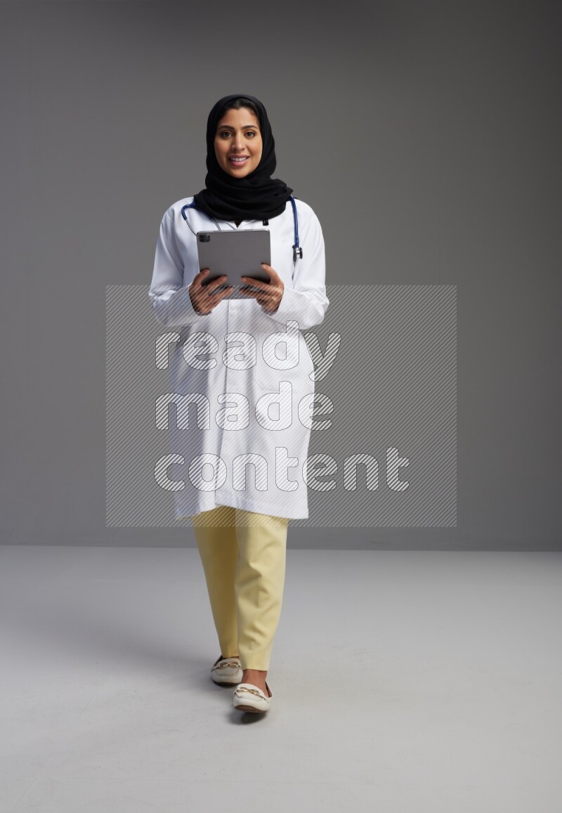 Saudi woman wearing lab coat with stethoscope standing working on tablet on Gray background