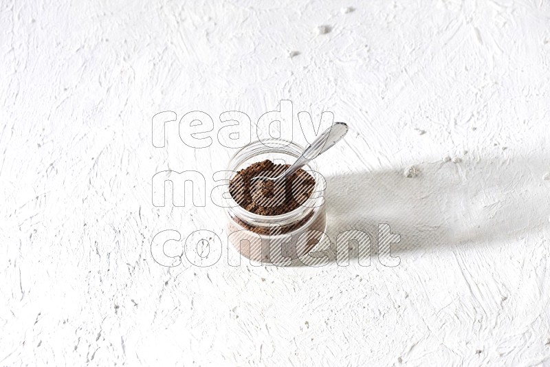 A glass jar full of cloves powder with a metal spoon on a textured white flooring