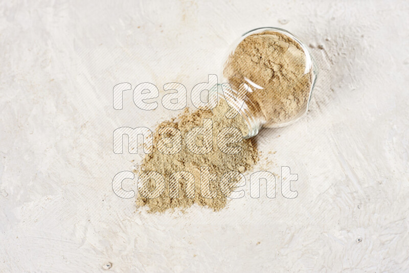 A glass jar full of ground ginger powder flipped with some spilling powder on white background