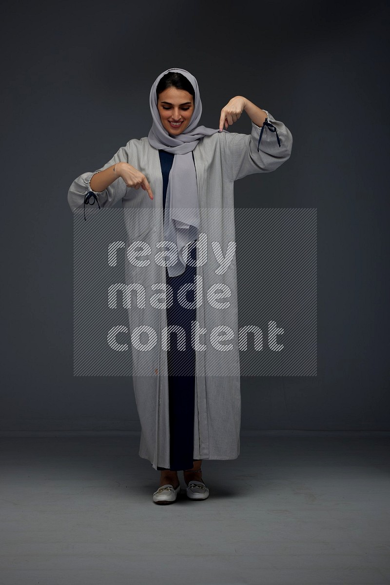 A Saudi woman wearing a light gray Abaya and head scarf standing and pointing in different directions eye level on a grey background