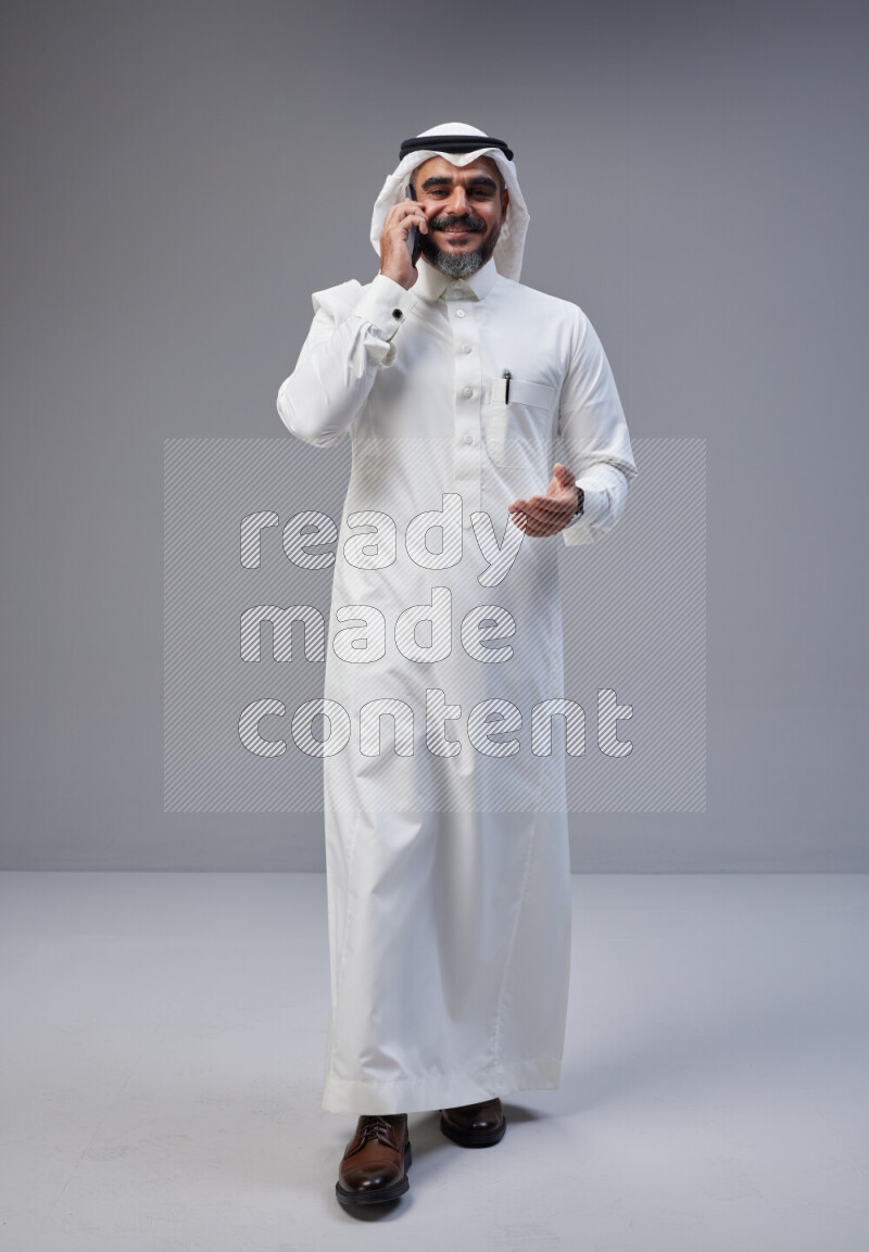 Saudi man Wearing Thob and white Shomag standing talking on phone on Gray background