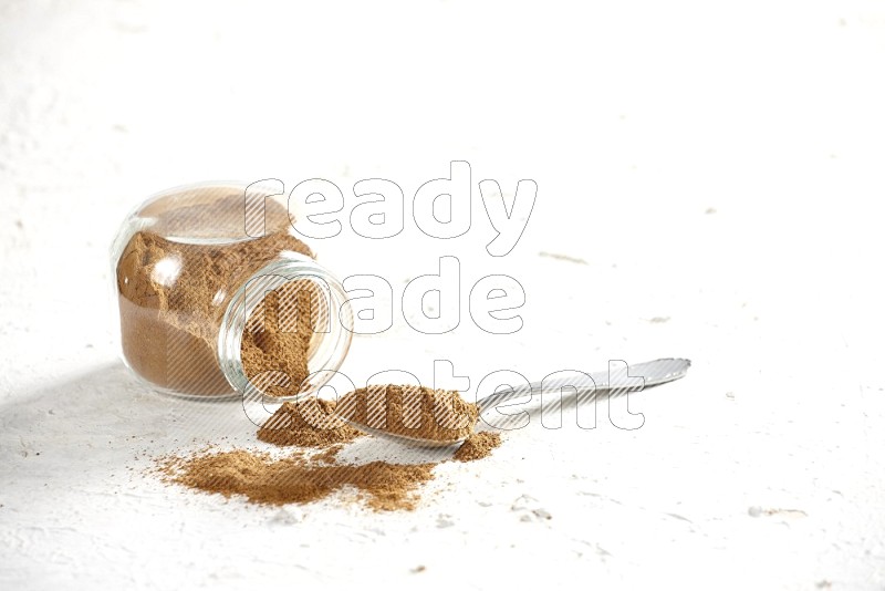 Flipped herbs glass jar full of cinnamon powder with a metal spoon full of powder on a textured white background