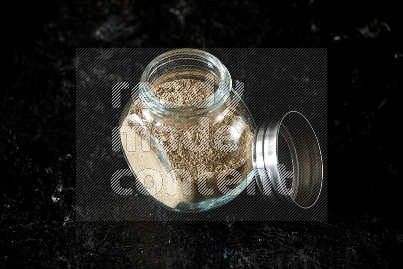 A glass spice jar full of cardamom powder on textured black flooring