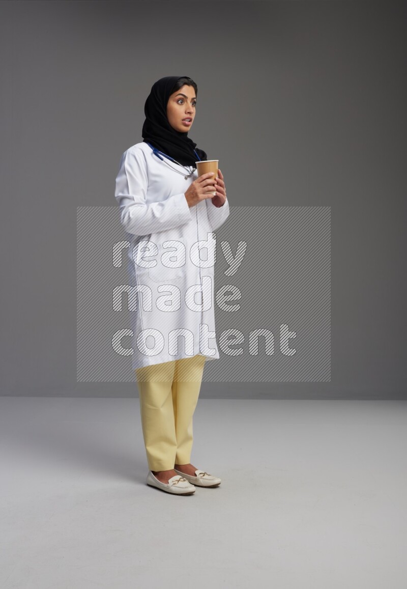 Saudi woman wearing lab coat with stethoscope standing holding paper cup on Gray background