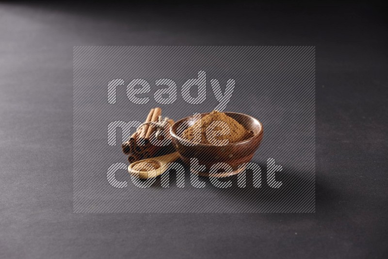 Cinnamon sticks stacked and bounded beside a wooden bowl full of cinnamon powder and a wooden spoon full of powder on black background