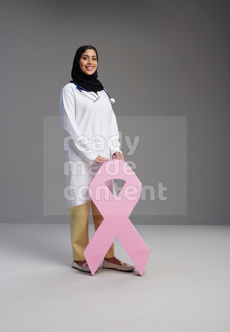 Saudi woman wearing lab coat with stethoscope standing holding awareness ribbon symbols standing on Gray background