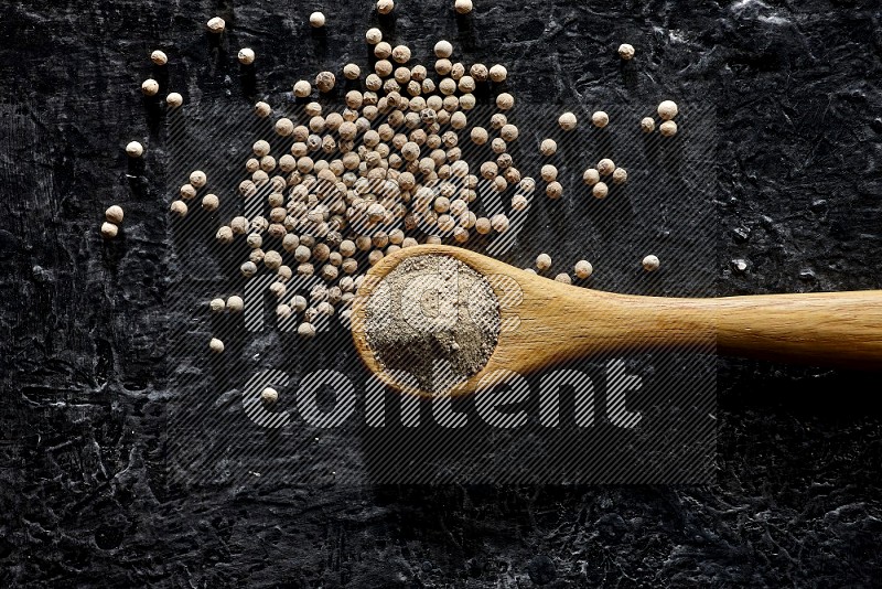 A wooden spoon full of white pepper powder with white pepper beads on textured black flooring