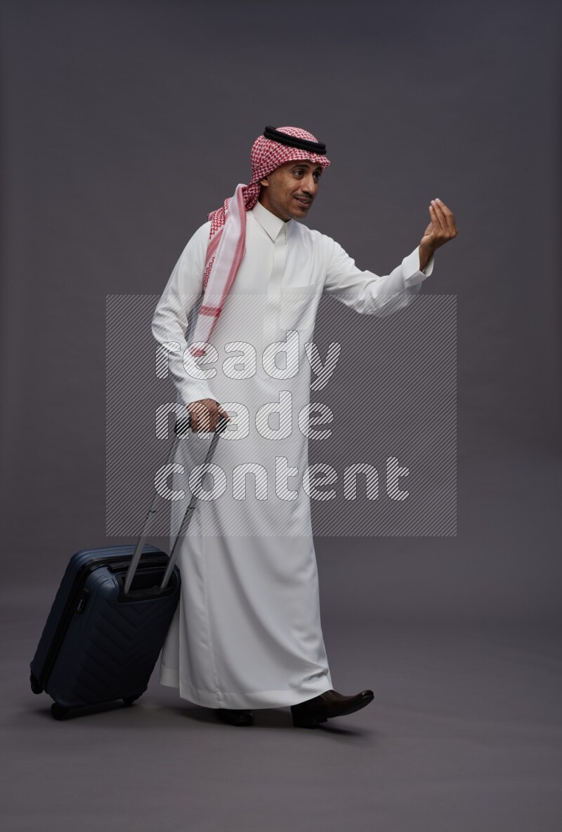 Saudi man wearing thob and shomag standing holding bag on gray background