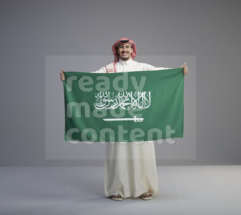 A saudi man standing wearing thob and red shomag holding big saudi flag on gray background