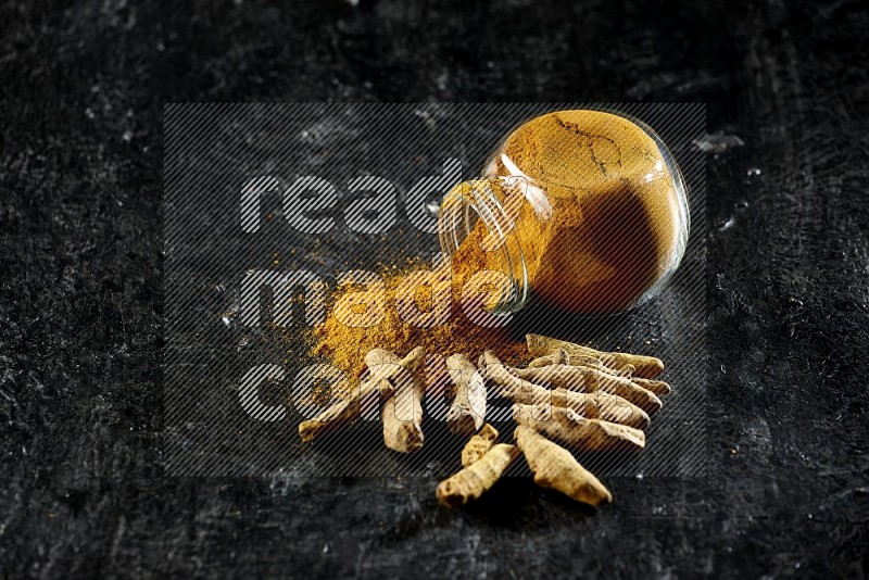 A flipped glass spice jar full of turmeric powder and powder spilled out of it with dried whole fingers on textured black flooring