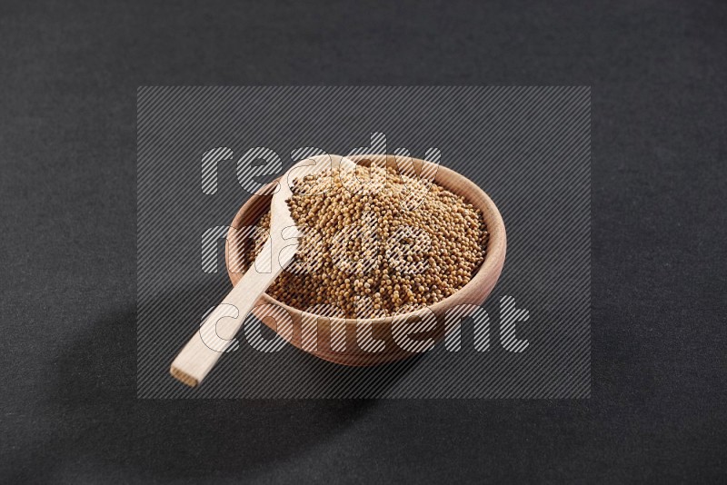 وعاء خشبي ممتلئ ببذور الخردل مع ملعقة خشبية ممتلئة فوق الوعاء علي خلفية سوداء
