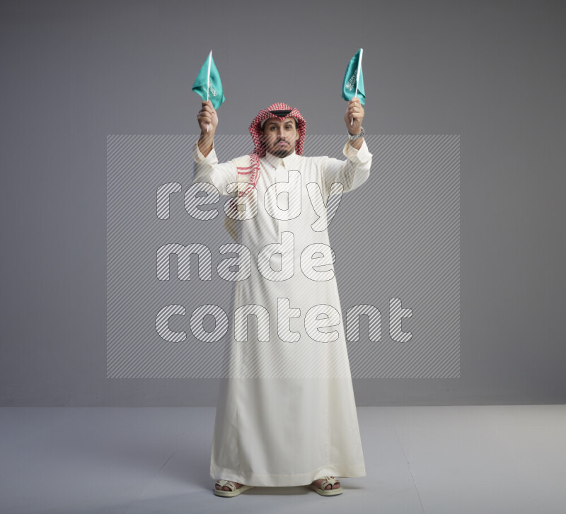 A Saudi man standing wearing thob and red shomag raising small Saudi flag on gray background