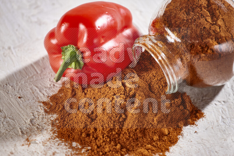 A glass jar full of ground paprika powder flipped with some spilling powder on white background