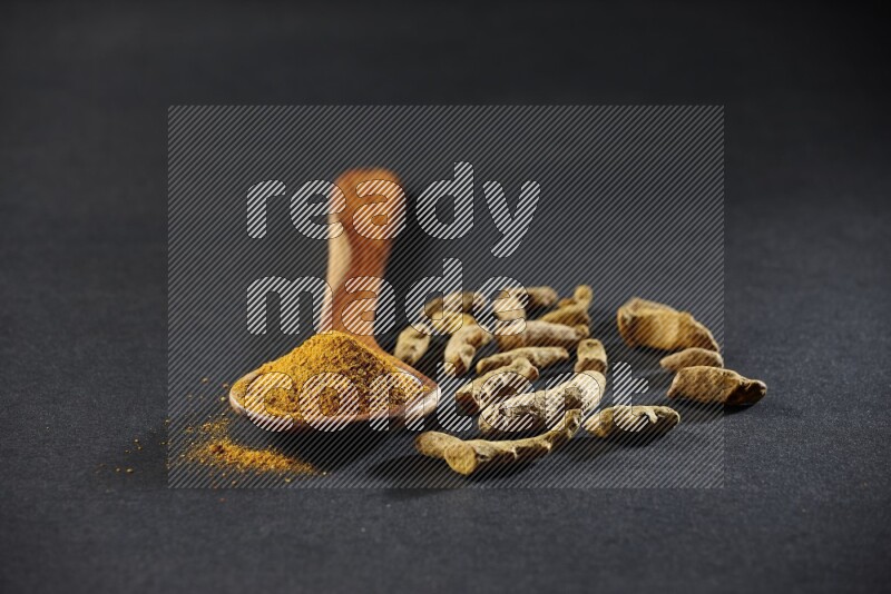 A wooden ladle full of turmeric powder with dried turmeric fingers on black flooring