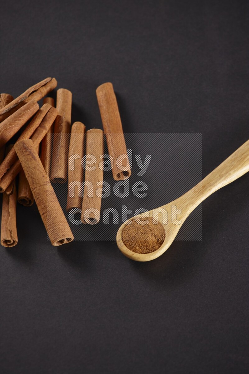 Cinnamon sticks stacked beside a wooden spoon full of cinnamon powder on black background