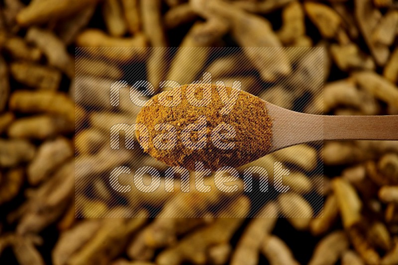 A wooden spoon full of turmeric powder above dried turmeric fingers on black flooring