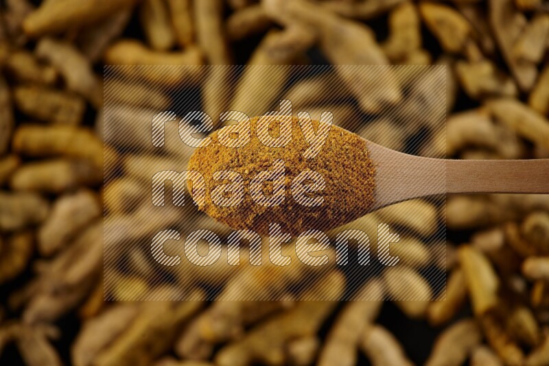 A wooden spoon full of turmeric powder above dried turmeric fingers on black flooring