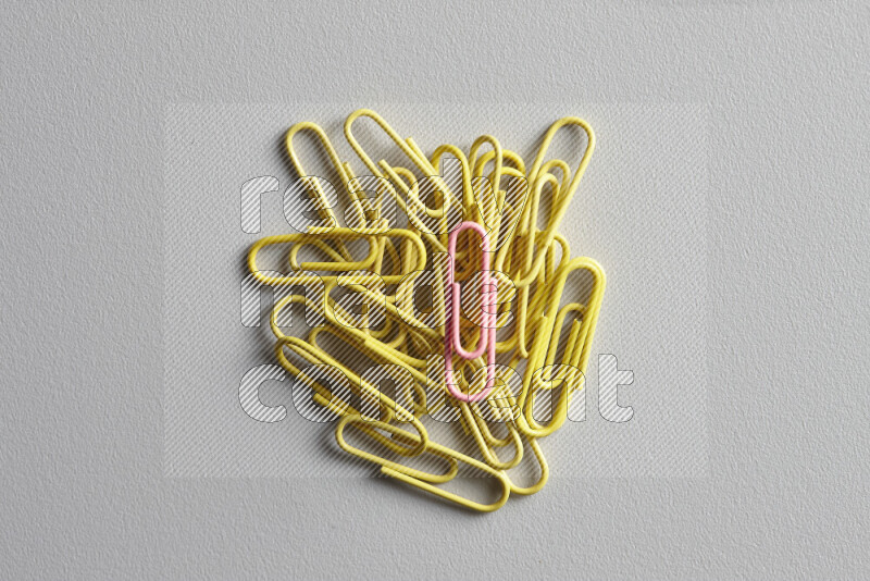 A pink paperclip surrounded by bunch of yellow paperclips on grey background