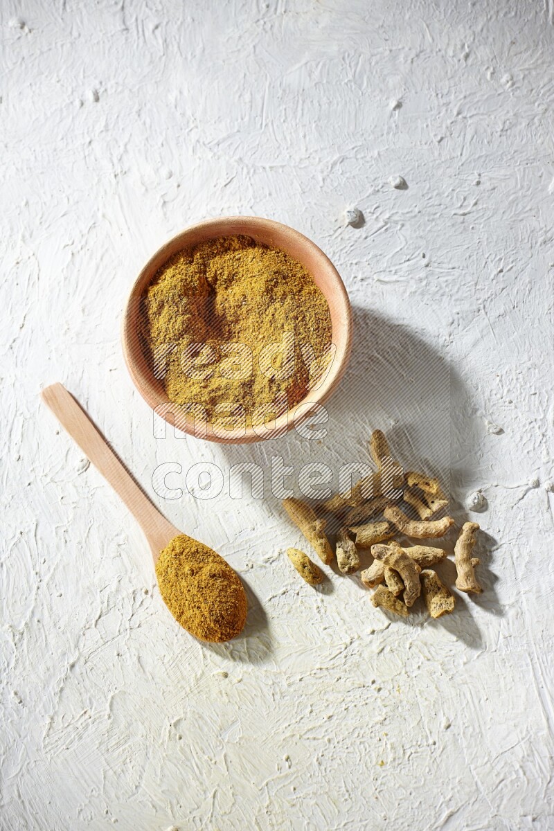 A wooden bowl and wooden spoon full of turmeric powder with dried turmeric fingers on textured white flooring